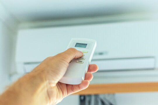 The hand turns on the air conditioner using the remote control. There is an air conditioner on the wall in the background.