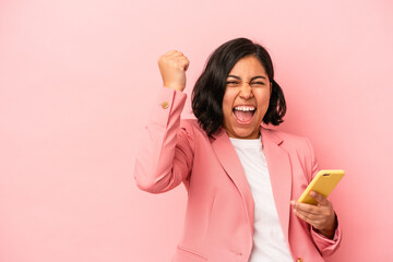 Young latin woman holding mobile phone isolated on pink background raising fist after a victory,...