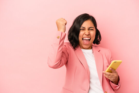 Young latin woman holding mobile phone isolated on pink background raising fist after a victory, winner concept.