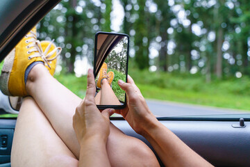 Close up with selective focus of unrecognizable woman using smartphone in a car. Horizontal view of woman with phone taking a picture of a forest from a car window . Technology and travel concept.