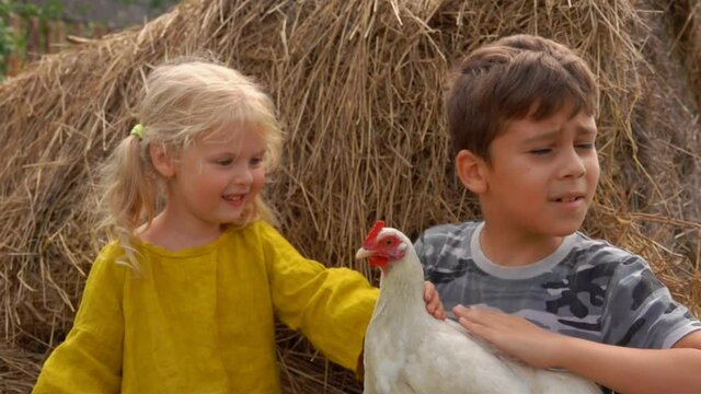Farmer boy and little girl are sitting on haystack and stroking a white chicken