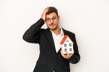 Young caucasian real estate holding a toy home isolated on white background being shocked, she has...