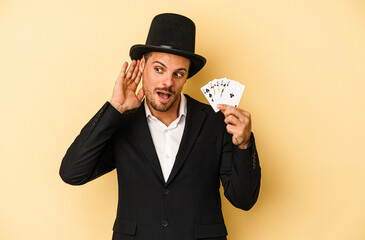 Young caucasian wizard holding magic card isolated on yellow background trying to listening a...
