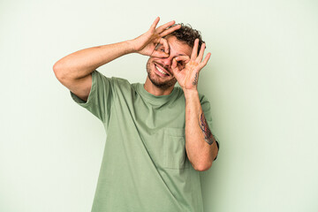 Young caucasian man isolated on green background showing okay sign over eyes