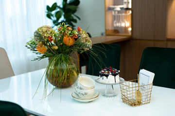 An exquisite berry cake on a glass stand on a table with a vase of flowers and a tea set in a modern designer interior. Soft selective focus, art noise