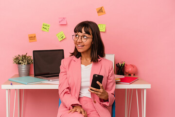 Young mixed race woman preparing a exam for university holding a mobile phone isolated on pink background looks aside smiling, cheerful and pleasant.