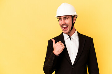 Young architect caucasian man isolated on yellow background points with thumb finger away, laughing and carefree.