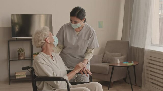 Medium stab shot of caring female social worker and old lady in wheelchair wearing face masks having conversation indoors