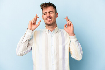 Young caucasian man isolated on blue background crossing fingers for having luck