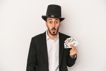 Young magician man holding a magic card isolated on white background shrugs shoulders and open eyes confused.