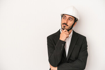 Young architect man wearing a construction helmet isolated on white background looking sideways with doubtful and skeptical expression.