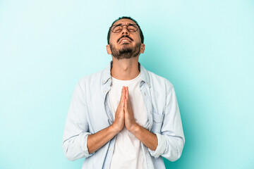 Young caucasian man isolated on blue background holding hands in pray near mouth, feels confident.