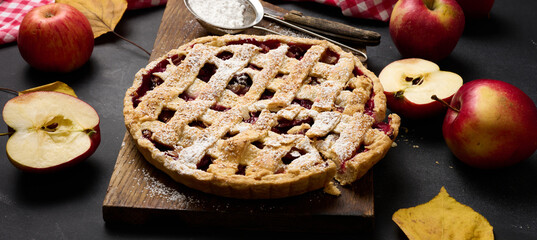 baked round traditional apple pie on brown wooden board and fresh red apples, top view