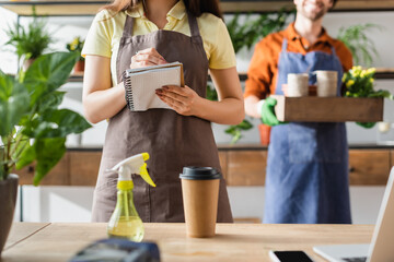 Cropped view of florist writing on notebook near coffee, devices and plant in flower shop