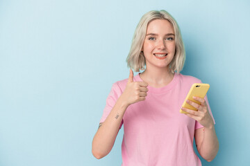 Young caucasian woman holding mobile phone isolated on blue background smiling and raising thumb up