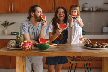A family in the kitchen cuting and eating the watermelon at home. Happy family of mother, father and lttle daughter eating fruits at home kitchen, have fun wit slices of watermelon