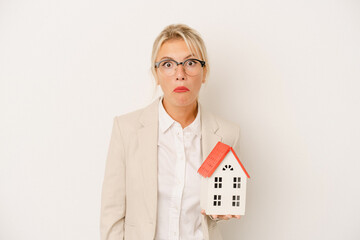 Young real estate agent woman holding a home model isolated on white background shrugs shoulders...