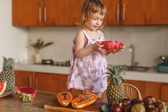 Daughter Helping Mother To Prepare Family Breakfast. Little girl stand by the table on the kitchen and hlding tropical dragon fruit in her hands