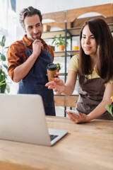 Seller in apron holding smartphone and pointing at laptop near colleague with paper cup in flower shop