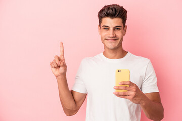 Young caucasian man holding a mobile phone isolated on pink background showing number one with finger.