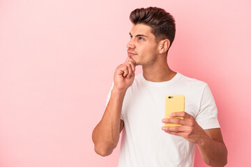 Young caucasian man holding a mobile phone isolated on pink background looking sideways with doubtful and skeptical expression.