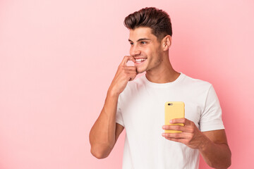 Young caucasian man holding a mobile phone isolated on pink background relaxed thinking about something looking at a copy space.