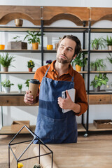 Seller in apron holding paper cup and notebook in flower shop