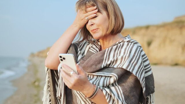 Handsome mature woman looking at phone and rubbing her forehead on the beach