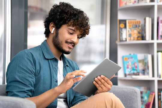 Young student in earphones using mobile tablet sitting on armchair in library