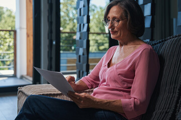 Attentive aging woman in glasses reading paper from armchair
