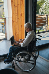 Male wheelchair user working on laptop near house window