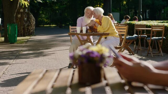 In a sunny day old beautiful couple with a large smile hugging each other in the park at cafe and discussing lovely together