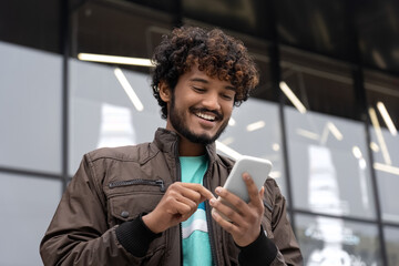 Happy young Indian man smiling holding mobile phone text social media outdoors
