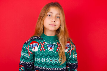 Happy brunette kid girl in knitted sweater christmas over red background looking at camera with charming cute smile.
