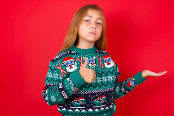 Happy cheerful brunette kid girl in knitted sweater Christmas over red background  showing thumb up and pointing with the other hand. I recommend this.