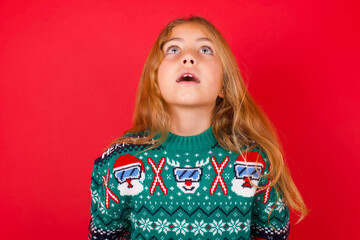Surprised brunette kid girl in knitted sweater Christmas over red background , shrugs shoulders, looking sideways, being happy and excited. Sudden reactions concept.