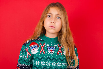 brunette kid girl in knitted sweater Christmas over red background  expressing disgust, unwillingness, disregard having tensive look frowning face, looking indignant with something.
