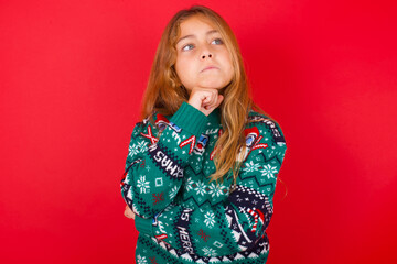 brunette kid girl in knitted sweater Christmas over red background  with hand under chin and looking sideways with doubtful and skeptical expression, suspect and doubt.