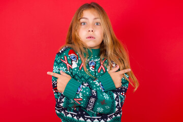 Confused brunette kid girl in knitted sweater Christmas over red background  chooses between two ways, points at both sides with crossed hands, feels doubt. Need your advice.