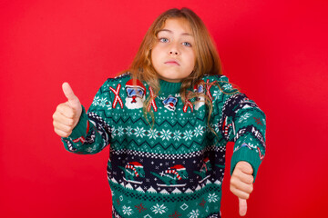 brunette kid girl in knitted sweater Christmas over red background  feeling unsure making good bad sign. Displeased and unimpressed.