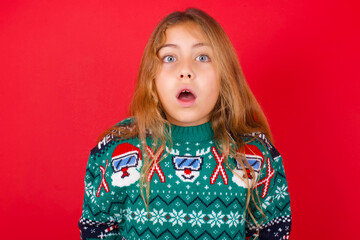 Emotional attractive brunette kid girl in knitted sweater Christmas over red wall with opened mouth expresses great surprise and fright, stares at camera. Unexpected shocking news and human reaction.