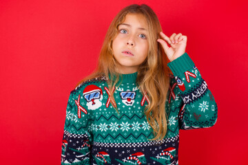 Shocked brunette kid girl in knitted sweater Christmas over red background  shows something little with hands, demonstrates size, opens mouth from surprise. Measurement concept.