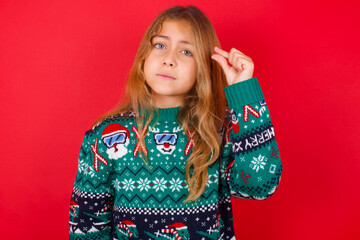Upset brunette kid girl in knitted sweater Christmas over red background  shapes little gesture with hand demonstrates something very tiny small size. Not very much