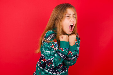 brunette kid girl in knitted sweater Christmas over red background  excited and glad to achieve victory, clenches fists, screams in excitement with closed eyes,successful person.