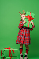 cute little girl of 7 years old with curly hair in golden deer horns, standing with gift boxes on a green background in the studio. the child smiles happily and looks away. Advertising. copy space