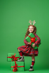 cute little girl of 7 years old with curly hair in golden deer horns, standing with gift boxes on a green background in studio. child smiles happily and looks into the frame. Advertising. copy space