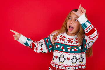 brunette kid girl in knitted sweater christmas over red background showing loser sign and pointing...