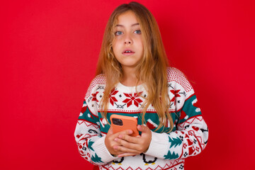 Obraz na płótnie Canvas Photo of astonished crazy brunette kid girl in knitted sweater christmas over red background hold smartphone dislike feedback concept
