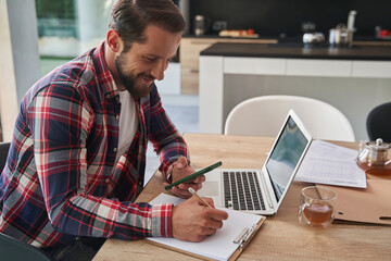 Bearded stylish male in shirt writing notes while holding mobile phone in room indoors