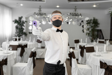 Waitress man wearing a face mask and holding an infrared thermometer on her forehead to check the...